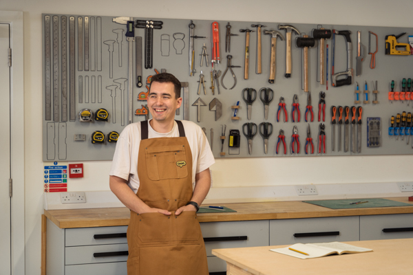 Design Manager Josh, stood in the workshop with tools behind him on the wall, wearing an Omlet work apron.