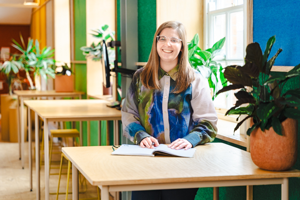 Rebecca, product design researcher stood at a wooden table with a notebook and plants, smiling at thr camera.